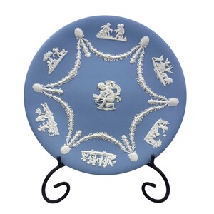 May include: A blue ceramic plate with white relief detailing, featuring cherubs and floral garlands. The plate is displayed on a black metal stand. The decorative plate has a classic, elegant design.