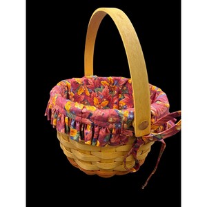 May include: A woven basket with a wooden handle and a floral fabric lining. The fabric features a pattern of red, orange, and yellow leaves. A matching fabric bow is tied on the side. The basket is designed for decorative or functional use.
