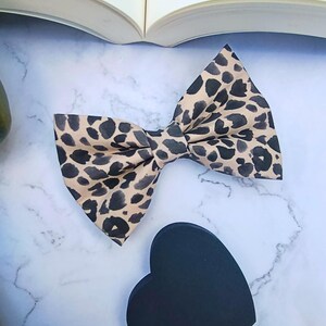 Peut inclure: Un nœud papillon à imprimé léopard beige et noir, accompagné d'un objet noir en forme de cœur. Le nœud papillon est en tissu et a une forme classique. Le fond est une surface en marbre avec un livre ouvert.