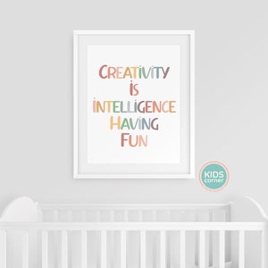 Op de afbeelding: Een witte ingelijste print met de tekst "CREATIVITY IS INTELLIGENCE HAVING FUN" in kleurrijke letters op een witte achtergrond. De print hangt aan een witte muur boven een wit babybedje.