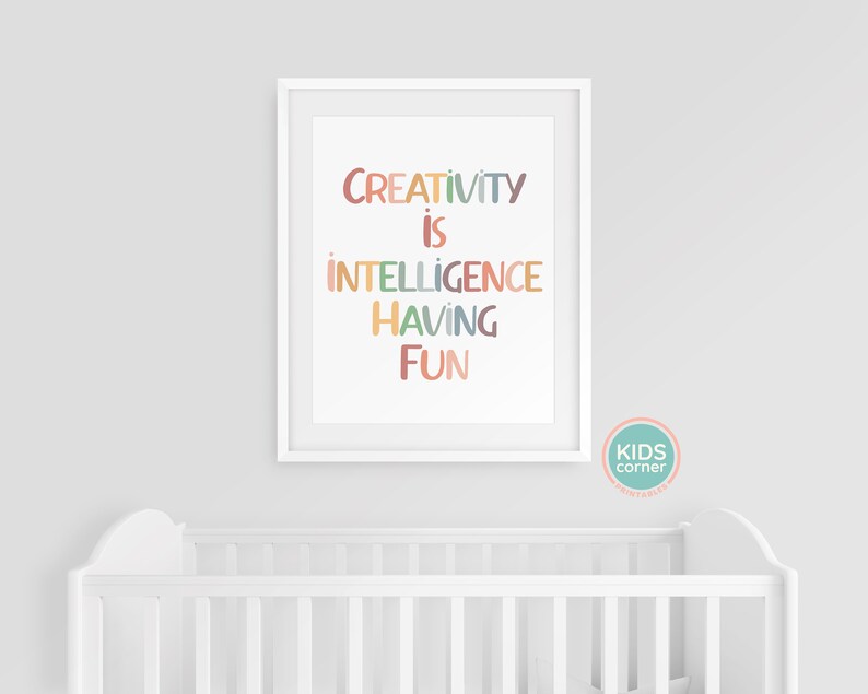 Op de afbeelding: Een witte ingelijste print met de tekst "CREATIVITY IS INTELLIGENCE HAVING FUN" in kleurrijke letters op een witte achtergrond. De print hangt aan een witte muur boven een wit babybedje.
