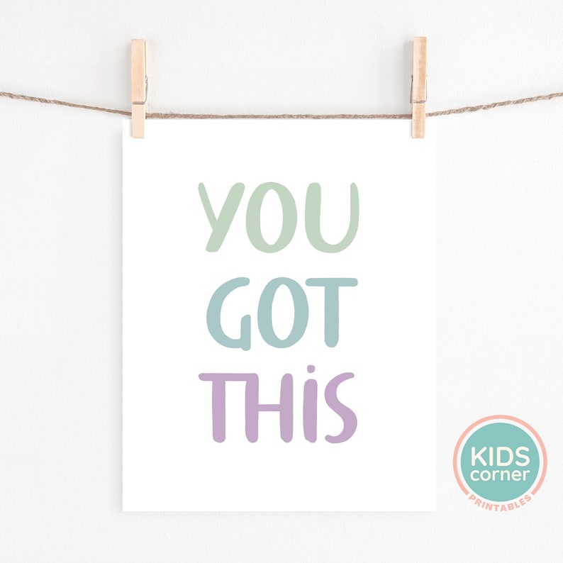 May include: A printable wall art print with the text "YOU GOT THIS" in pastel green, blue, and purple colors. The print is hanging on a white wall with a string and wooden clothespins.