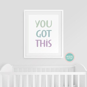 May include: White framed print with the text "YOU GOT THIS" in pastel green, blue, and purple lettering. The print is hanging on a white wall above a white crib.