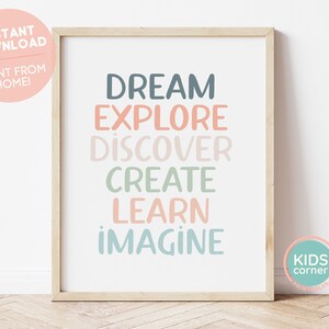 May include: A printable wall art print with the words "Dream, Explore, Discover, Create, Learn, Imagine" in a colorful, layered font style. The print is framed in a light wood frame and is set against a white wall.