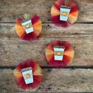 May include: Four fall-themed hair clips. Each clip features a glittery coffee cup with a pumpkin design, set against a backdrop of layered tulle in shades of red, orange, and yellow. The clips are arranged on a wooden surface.