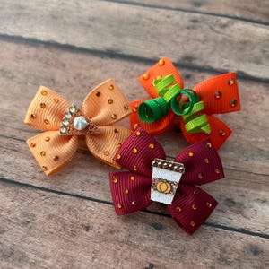 May include: Three hair bows made of ribbed fabric with small orange rhinestones. One light orange bow features a slice of pie, one bright orange bow has green curly ribbons, and one dark red bow displays a coffee cup with a pumpkin icon.