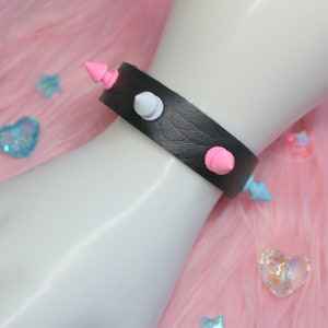 Peut inclure: Un bracelet en cuir noir avec trois pointes, une blanche, une rose et une bleue.