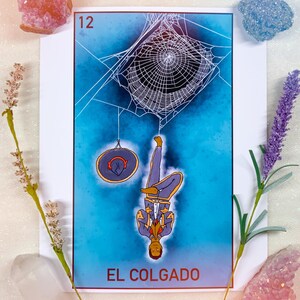 May include: A tarot card labeled 12 and EL COLGADO featuring a figure hanging upside down from a spider web, accompanied by a circular object. The card is surrounded by a pink crystal, a blue crystal, a clear crystal, and two sprigs of lavender.