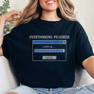 Könnte beinhalten: Schwarzes T-Shirt mit dem Text "OVERTHINKING PROGRESS" über einer Computer-Ladefenstergrafik. Die Grafik zeigt einen blauen Fortschrittsbalken und das Wort "Loading...". Das Shirt wird mit einer blauen Jeans kombiniert.