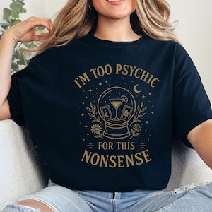 Könnte beinhalten: Schwarzes T-Shirt mit goldfarbener Grafik einer Kristallkugel, Mond und floralen Elementen. Der Text lautet "I'M TOO PSYCHIC FOR THIS NONSENSE". Das Shirt wird mit einer blauen Jeans getragen.