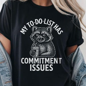 Op de afbeelding: Zwart T-shirt met een afbeelding van een wasbeer die een drankje vasthoudt, en de witte tekst "MY TO-DO LIST HAS COMMITMENT ISSUES". De wasbeer is in zwart-wit. Het shirt heeft een casual stijl.