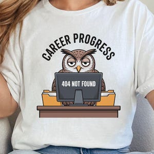 May include: White t-shirt with a cartoon owl design. The owl is sitting at a desk with a computer screen displaying "404 NOT FOUND". The text "CAREER PROGRESS" is arched above the owl. The shirt is worn with blue jeans.
