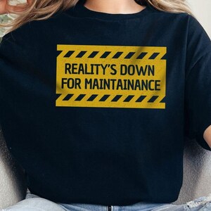 Könnte beinhalten: Schwarzes T-Shirt mit dem Text "REALITY'S DOWN FOR MAINTAINANCE" in einem gelb-schwarzen Warnschild-Design. Die Person trägt zerrissene blaue Jeans.