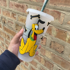Puede incluir: Un vaso de plástico transparente con una imagen de dibujos animados de Pluto el perro. El vaso tiene un diseño de hueso blanco y negro y una pajita verde.