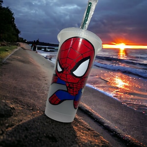 Puede incluir: Una taza reutilizable blanca con un dise&ntilde;o de Spiderman rojo y azul. La taza est&aacute; en una playa de arena con una puesta de sol en el fondo.