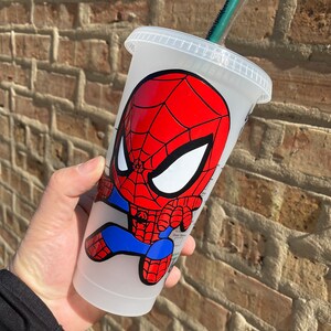 Puede incluir: Un vaso reutilizable de pl&aacute;stico transparente con una imagen de dibujos animados de Spiderman en rojo y azul. El vaso tiene una tapa negra y una pajita verde.