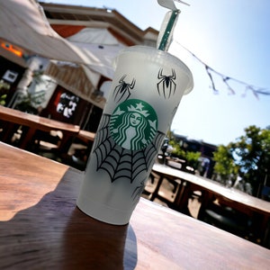 Puede incluir: Un vaso reutilizable de pl&aacute;stico transparente con un dise&ntilde;o de telara&ntilde;a en blanco y negro, un logotipo de Starbucks verde y dos ara&ntilde;as negras. El vaso est&aacute; sobre una mesa de madera.