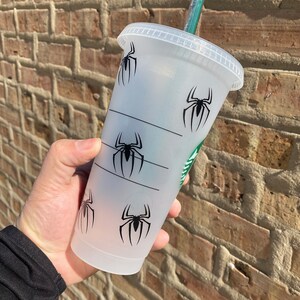 Puede incluir: Un vaso reutilizable de pl&aacute;stico transparente con un dise&ntilde;o gr&aacute;fico de ara&ntilde;a negra. El vaso tiene una tapa negra y una pajita verde.