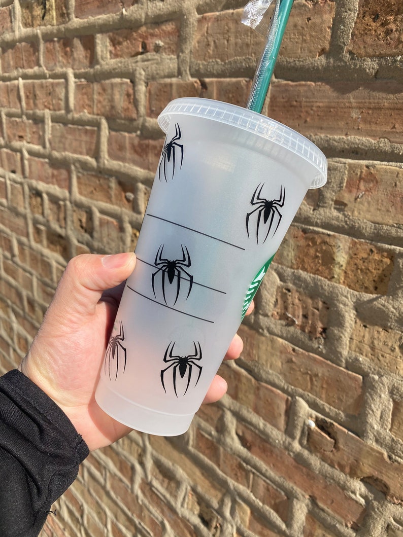 Puede incluir: Un vaso reutilizable de pl&aacute;stico transparente con un dise&ntilde;o gr&aacute;fico de ara&ntilde;a negra. El vaso tiene una tapa negra y una pajita verde.