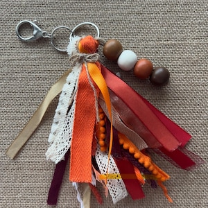 Op de afbeelding: Een sleutelhanger met een zilverkleurige karabijnhaak en ringen, bevestigd aan een kwast van diverse linten in tinten oranje, rood, bordeaux en crème, geaccentueerd met witte en bruine houten kralen en een klein stukje touw aan de bovenkant.