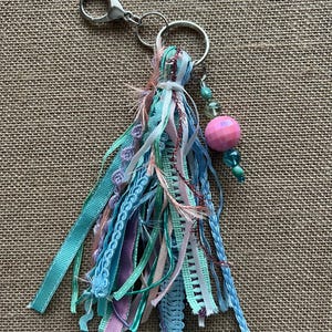 May include: A handmade keychain with a tassel design. The tassel is made of various ribbons in shades of blue, teal, pink, and purple. A pink beaded ball and turquoise beads are attached to the keychain. The keychain has a silver clasp and ring.