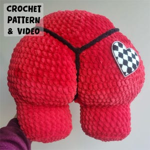 Peut inclure: Un grand coussin en crochet rouge en forme de cœur, avec une bordure noire et un patch en forme de cœur à carreaux. Le coussin a une surface texturée. Le texte dans le coin supérieur gauche indique "CROCHET PATTERN & VIDEO".