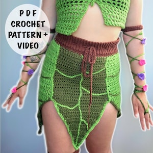 May include: A green crochet skirt and top set with a leaf-like design. The skirt has a brown waistband and a drawstring. The arm wraps are adorned with pink and purple flowers. The text "PDF CROCHET PATTERN + VIDEO" is in a white circle.