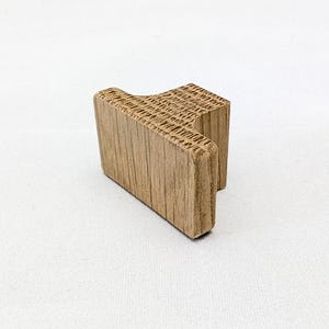 May include: A small, light brown wooden block in a distinctive T-shape. The wood grain is visible, and the block appears to be a decorative or functional item. The object is set against a plain white background.