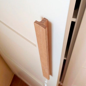 May include: A wooden cabinet door handle with a simple, rectangular design. The handle is made of light brown wood and is attached to a white cabinet door.