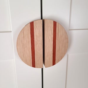 Peut inclure: Une paire de poignées de porte de placard en bois avec une forme arrondie en demi-cercle. Les poignées sont en bois clair avec deux bandes rouges qui descendent verticalement au centre de chaque poignée.