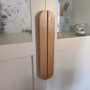May include: A pair of light oak cabinet pulls, mounted vertically on a white cabinet door. The pulls have a rounded top and a split design. The cabinet door is part of a white cabinet with glass doors, and floral wallpaper is visible inside.