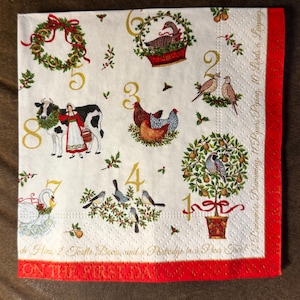 May include: A white Christmas-themed paper napkin with a red border. The napkin features illustrations of the twelve days of Christmas, including birds, a cow, and a pear tree. Text includes "On the first day" and "12 Drummers Drumming."