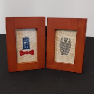 May include: Two small wooden picture frames with cross-stitch designs. The left frame shows a blue police box with a red bow tie. The right frame shows a gray Weeping Angel.