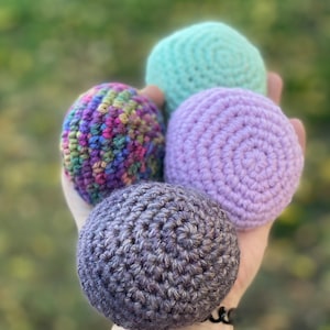 Puede incluir: Cuatro bolas de ganchillo coloridas en tonos morados, verdes y un hilo multicolor. Las bolas se sostienen en una mano, mostrando la textura y la calidad artesanal. Adecuado para malabares o como elementos decorativos.