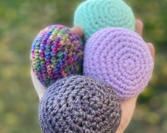 Könnte beinhalten: Vier farbenfrohe Häkelbälle in Lila, Grün und einem bunten Garn. Die Bälle werden in einer Hand gehalten, wodurch die Textur und die handwerkliche Qualität hervorgehoben werden. Geeignet zum Jonglieren oder als Dekoration.