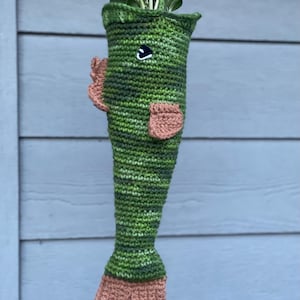 May include: A handmade, green crochet fish-shaped hanging planter. The planter features a detailed design with fins and a tail in shades of green and tan. The fish planter is filled with green leafy plants, adding a touch of nature to any space.