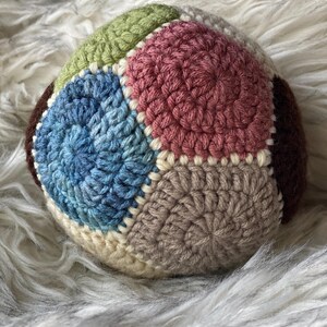 May include: A colorful, crocheted ball featuring a patchwork design. The ball is composed of various yarn colors, including blue, green, pink, brown, and beige. The handmade ball is likely a toy or decorative item.