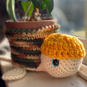 May include: A crocheted turtle toy wearing a yellow hat. The turtle has a white body and a blue eye. The turtle is sitting on a windowsill next to a potted plant.