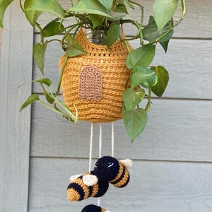 Puede incluir: Un macetero de crochet en forma de colmena con una puerta marrón y tres abejas de crochet colgando de la parte inferior. El macetero es amarillo y las abejas son negras y amarillas.