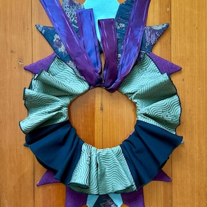 May include: A green, black, and purple fabric collar with a ruffled edge and a starburst design. The collar is made of different fabrics and has a ribbon tied at the top.