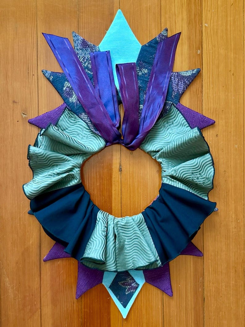 May include: A green, black, and purple fabric collar with a ruffled edge and a starburst design. The collar is made of different fabrics and has a ribbon tied at the top.
