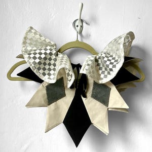 May include: A white and black checkered fabric collar with a starburst design. The collar is made of a combination of white, black, and gray fabric. It is hanging on a wooden hanger.