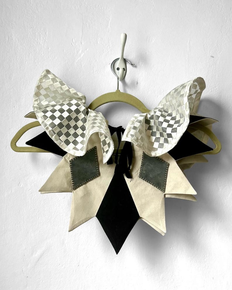 May include: A white and black checkered fabric collar with a starburst design. The collar is made of a combination of white, black, and gray fabric. It is hanging on a wooden hanger.