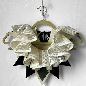 May include: A white ruffled collar with a checkered pattern, attached to a black fabric base with a green patch. The collar is hanging on a wooden hanger.