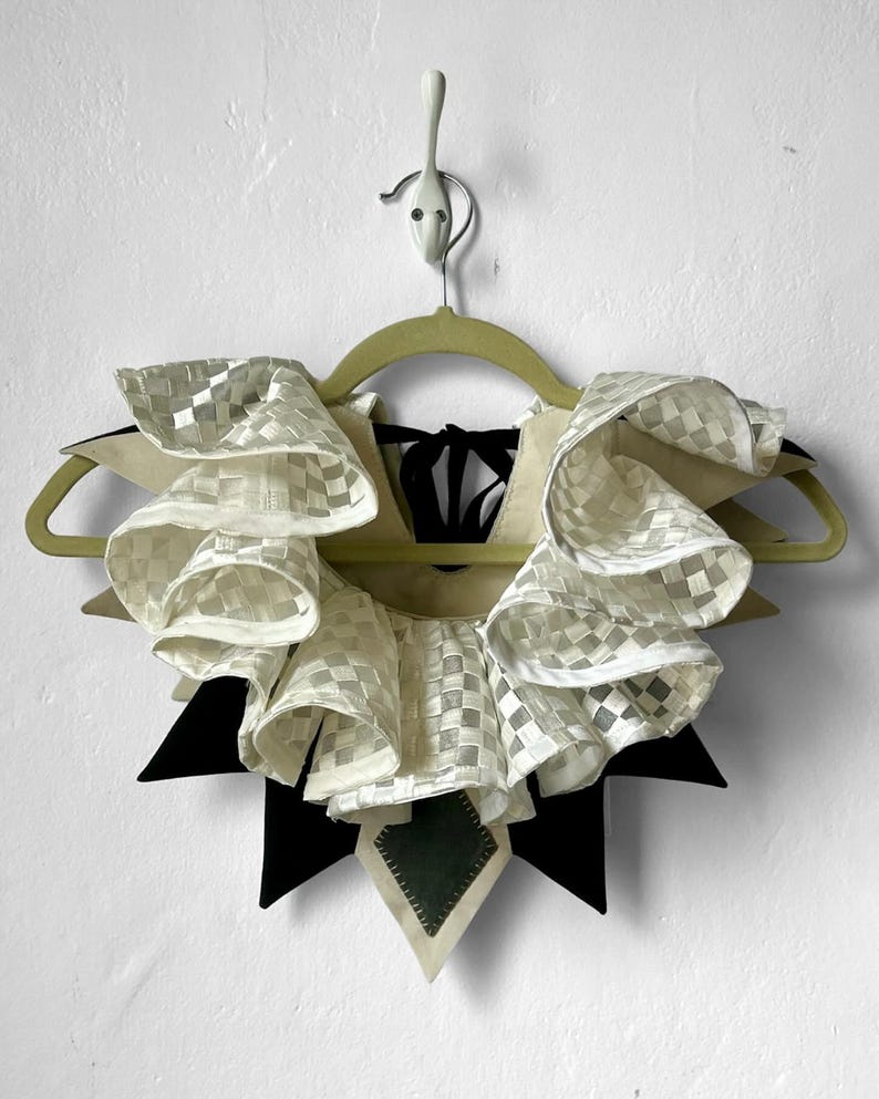 May include: A white ruffled collar with a checkered pattern, attached to a black fabric base with a green patch. The collar is hanging on a wooden hanger.