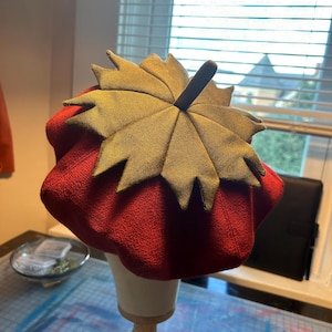 May include: A red beret with a green leaf-shaped top. The beret is on a wooden stand.