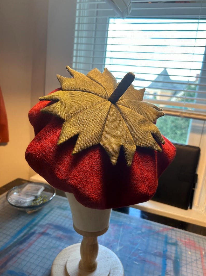 May include: A red beret with a green leaf-shaped top. The beret is on a wooden stand.