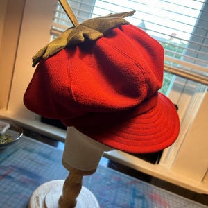 May include: A red felt newsboy cap with a green felt leaf on top. The cap is on a wooden display stand.