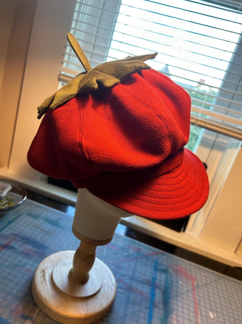 May include: A red felt newsboy cap with a green felt leaf on top. The cap is on a wooden display stand.