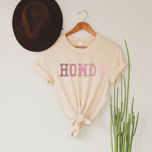 May include: A beige t-shirt with the word "HOWDY" printed in pink and white letters. The shirt is tied in a knot at the bottom.